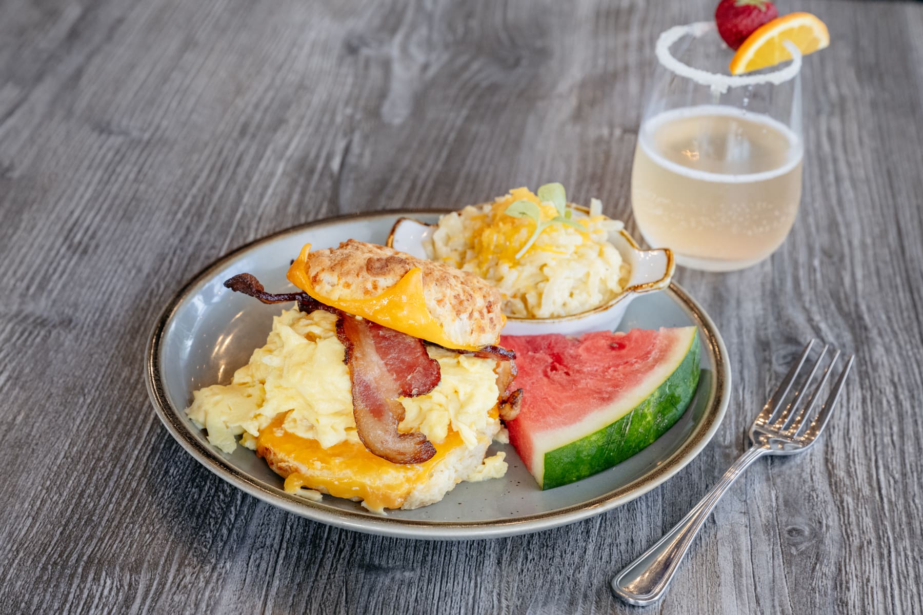 A bacon, egg and cheese biscuit on a plate with a slice of watermelon and cheesy potatoes, a sparkling drink is on the table beside the plate