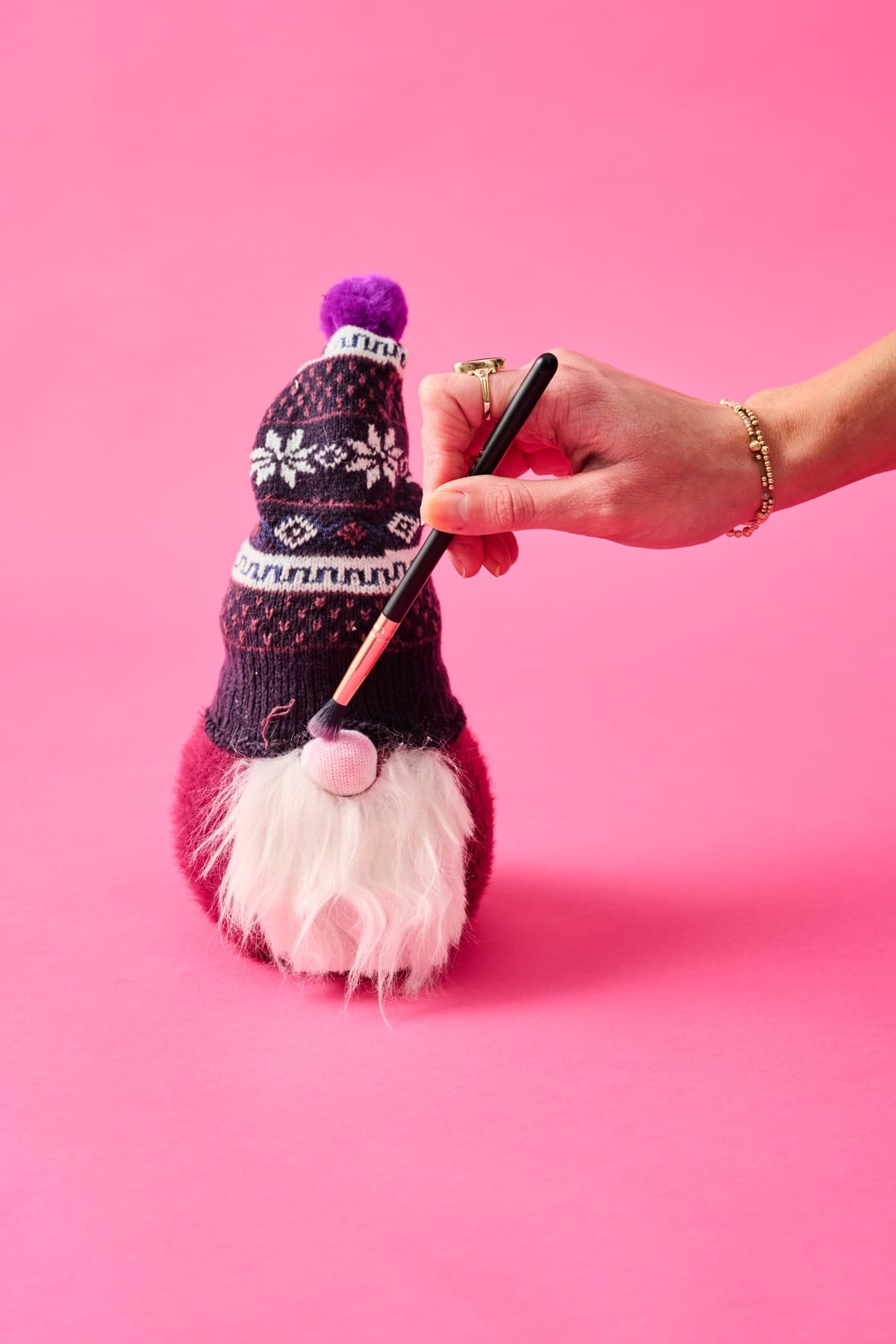 A hand reaching with a makeup brush to apply pink makeup to the nose of a sock gnome