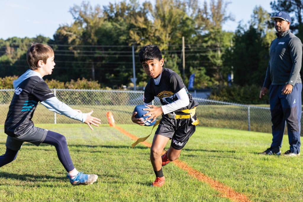 Youth flag football players in action while their coach oversees.
