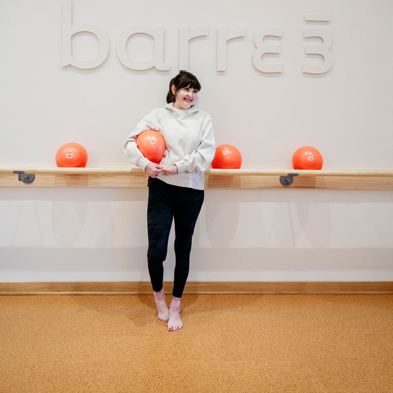Stephanie Nielson stands inside her workout studio Barre3 in workout clothes, holding a bright orange exercise ball with her studio's logo on it. A bar on the wall behind her holds up other orange exercise balls, and above that, her studio's name, "Barre3," is in large letters on the wall behind her.