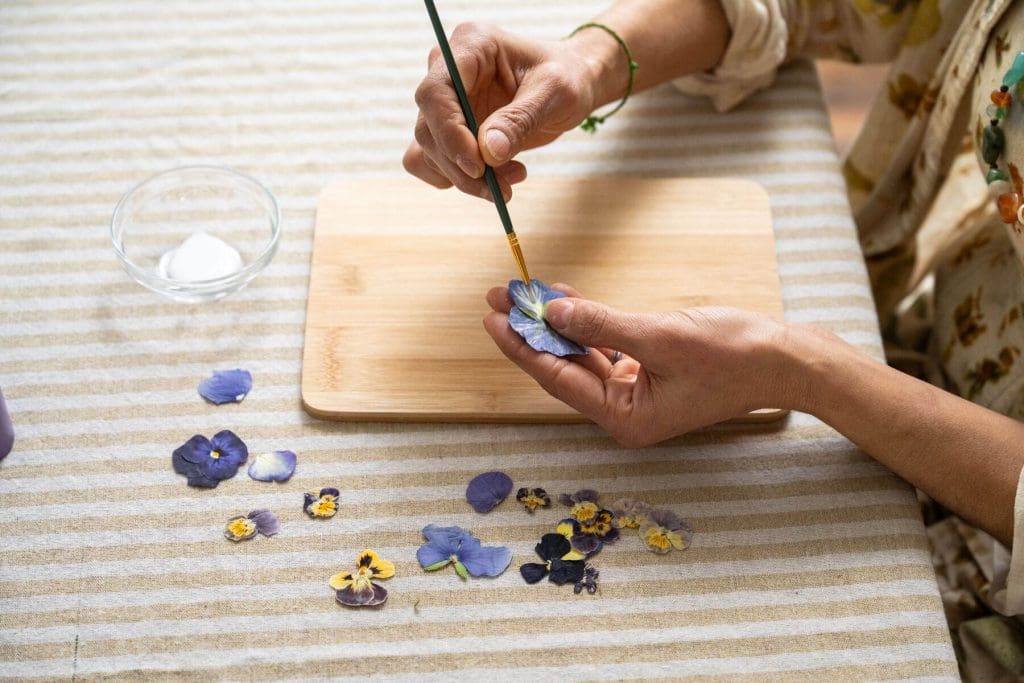 Deena Class using a paintbrush to glue a pressed pansy flower onto a craft surface for a botanical art project.