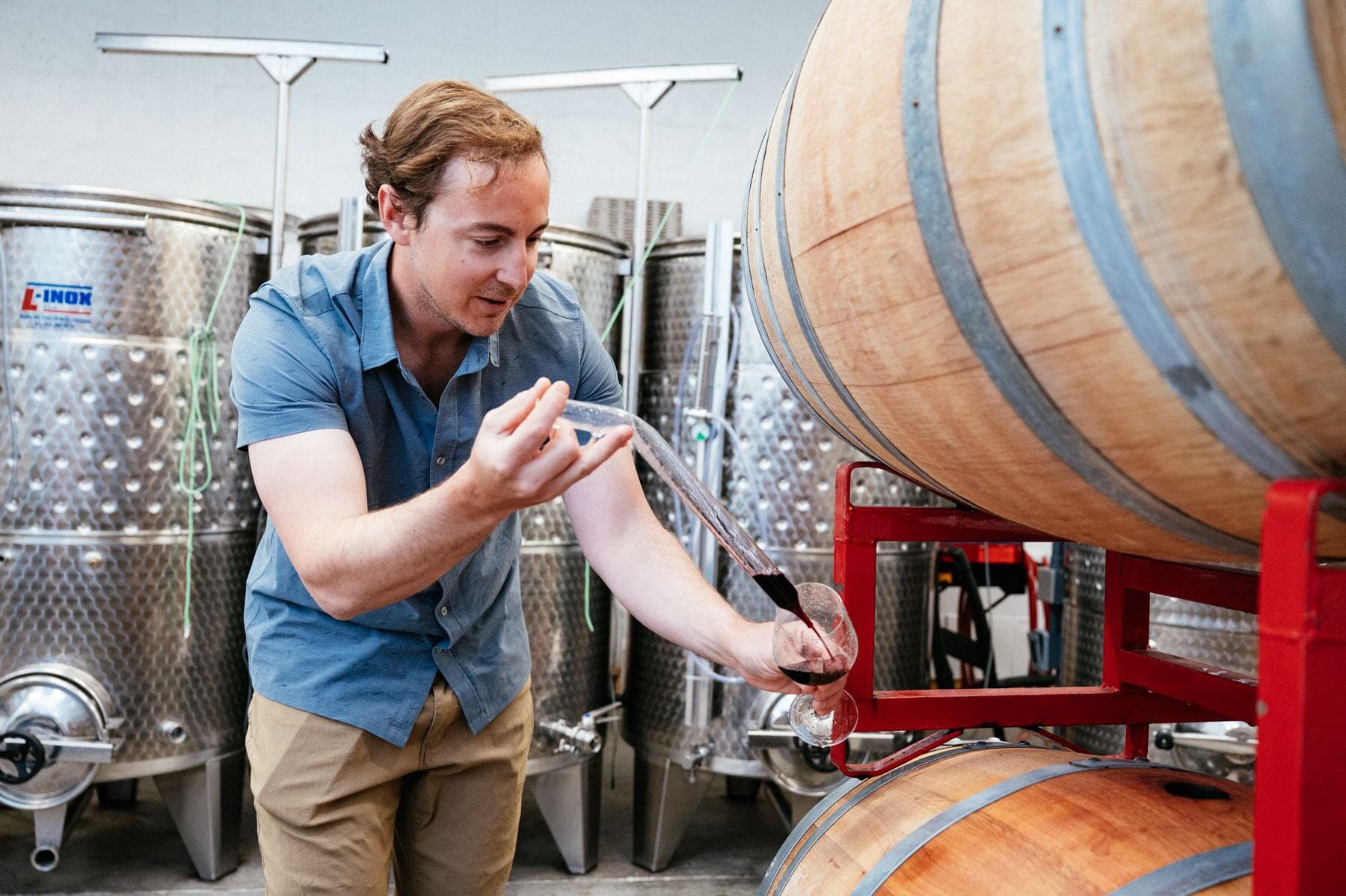 A man siphons red wine into a glass while standing next to a barrel. 