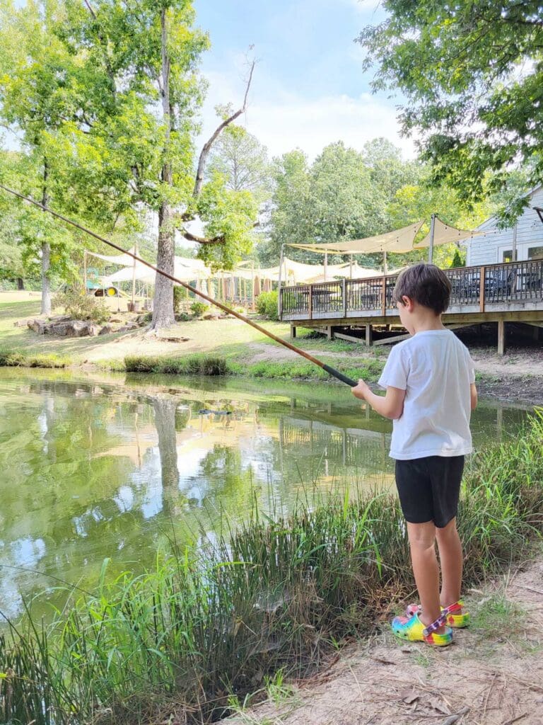 A kid stands outside fishing in a pond.