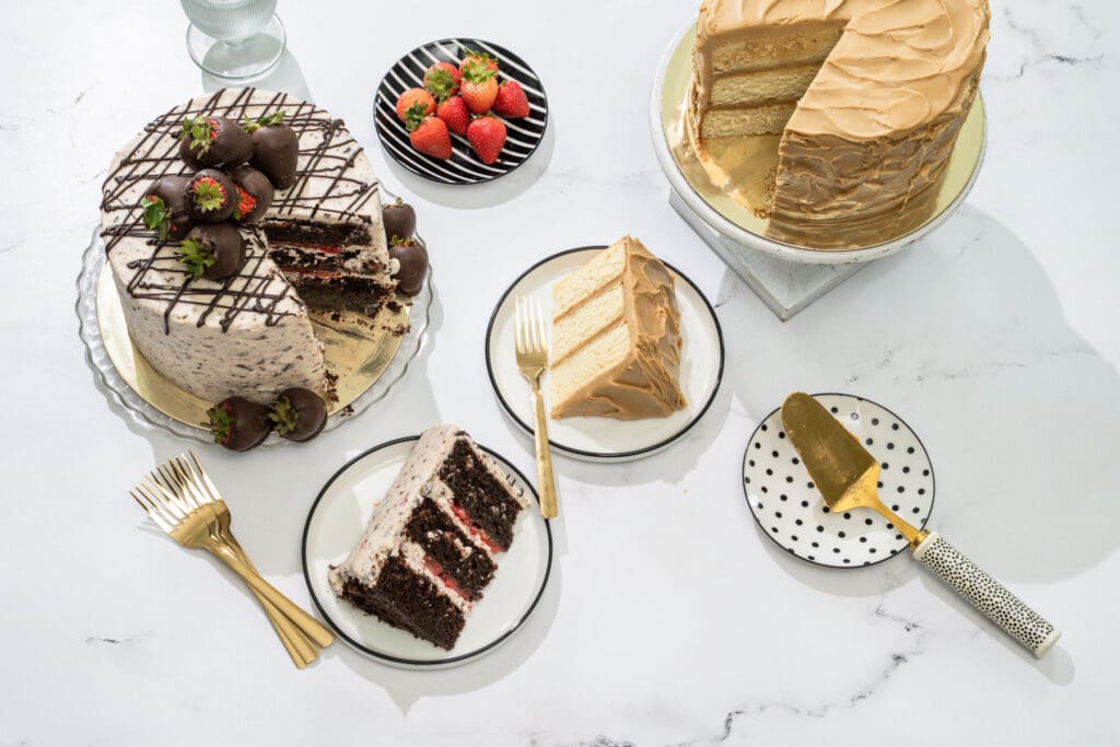 Overhead view of two sliced layer cakes on marble — a chocolate strawberry cake with chocolate-dipped berries and a caramel-frosted yellow cake — with plated slices, gold forks and a cake server
