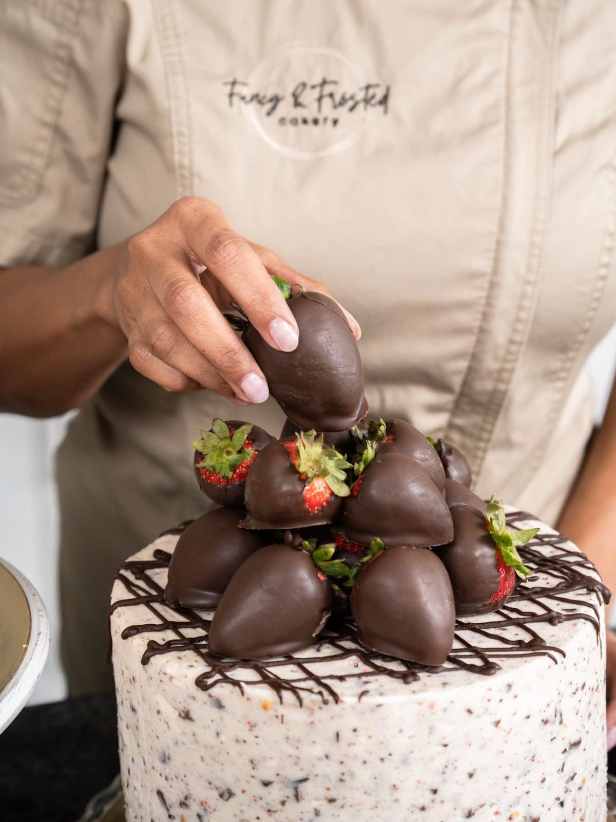 Hands in a tan Fancy & Frosted chef's coat stacking chocolate-covered strawberries on top of a speckled buttercream cake drizzled with chocolate.